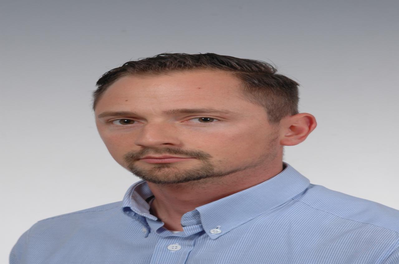 Portret mężczyzny w błękitnej koszuli z krótką brodą i ciemnymi włosami na jasnym tle.