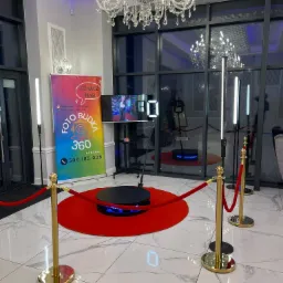 Wnętrze z gotową do użycia platformą obrotową 360 do nagrywania filmów, otoczoną czerwonym dywanem i odgrodzoną ozdobnymi słupkami z czerwonym sznurem, w tle widoczny ekran z nagrywanym filmem...