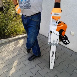 Mężczyzna w jeansach i koszulce z Popeye trzymający piłę spalinową Stihl, stojący na kostce brukowej przed jasną ścianą.