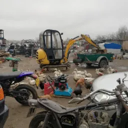 Mała koparka z łyżką załadowuje przyczepkę na podwórku pełnym złomu, gdzie spacerują kury i kaczki, a w pierwszym planie widać motocykle i samochody.