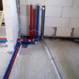 Surowe wnętrze z widocznymi, nowymi rurami instalacji wodnej (czerwone i niebieskie) oraz kanalizacyjnej (szare) wychodzącymi ze ściany z betonu komórkowego, zabezpieczone obejmami.