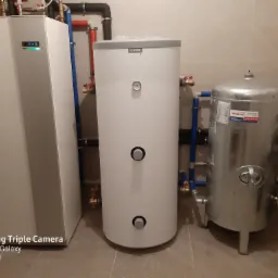 Zestaw urządzeń grzewczych w kotłowni: pompa ciepła Nibe, biały zasobnik wody Nibe z termometrem i srebrny zbiornik hydroforowy Wimest ZCS/300/6, połączone rurami w kolorach niebieskim, czerwonym...