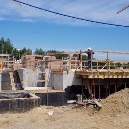 Izol-Mont Sp. z o.o. - Budowa domu w trakcie realizacji: widoczne fundamenty z izolacją, szalunki, dźwigi w tle i pracownik w kasku.