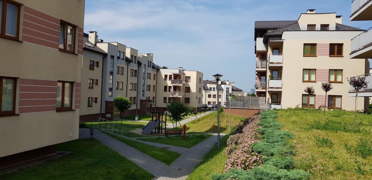 Kompleks budynków mieszkalnych z placem zabaw i zagospodarowaną zielenią, widok z perspektywy lekko podwyższonej, w tle błękitne niebo.