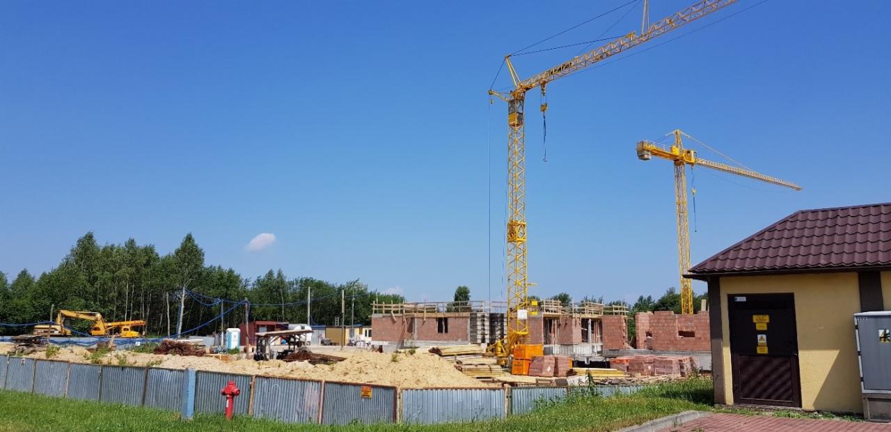 Budowa domu z cegły w trakcie realizacji, widoczne dwa żółte żurawie, ogrodzenie z blachy falistej oraz budynek gospodarczy z brązowym dachem.