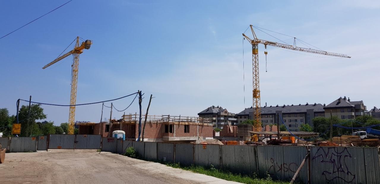 Budowa dwóch budynków z czerwonej cegły, widoczne dwa żółte dźwigi wieżowe na tle błękitnego nieba i istniejącego osiedla mieszkaniowego.
