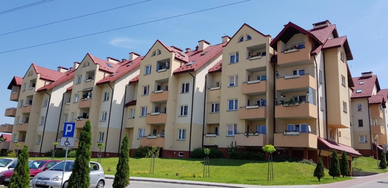 Nowoczesny, wielorodzinny budynek mieszkalny z czerwonym dachem i balkonami, otoczony zielenią i miejscami parkingowymi.