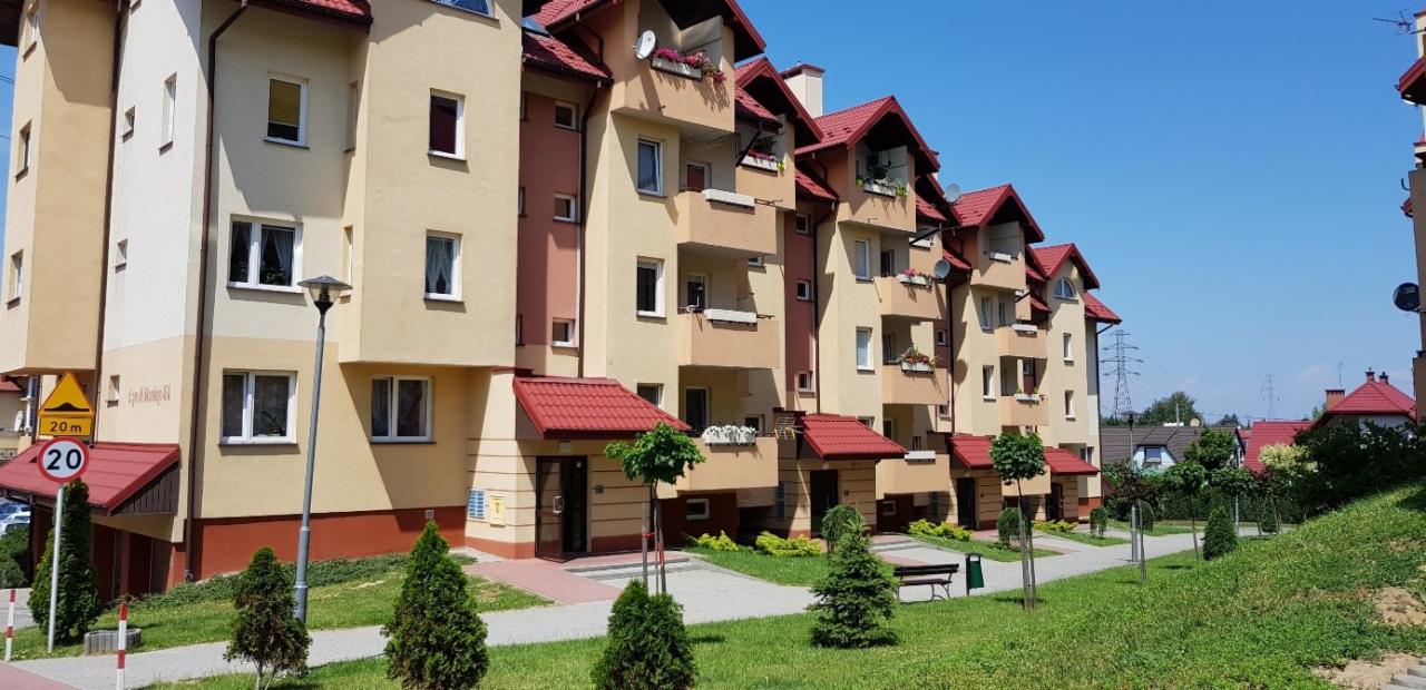 Nowoczesne, wielorodzinne budynki z jasną elewacją i czerwonymi dachami, z balkonami ozdobionymi kwiatami, widoczne znaki drogowe i zadbana zieleń.