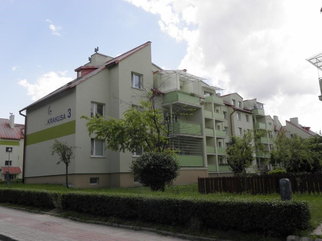 Wielopiętrowy budynek mieszkalny z balkonami, otynkowany na jasnozielono, z napisem 'Krakusa 3' na bocznej ścianie, otoczony zielenią.