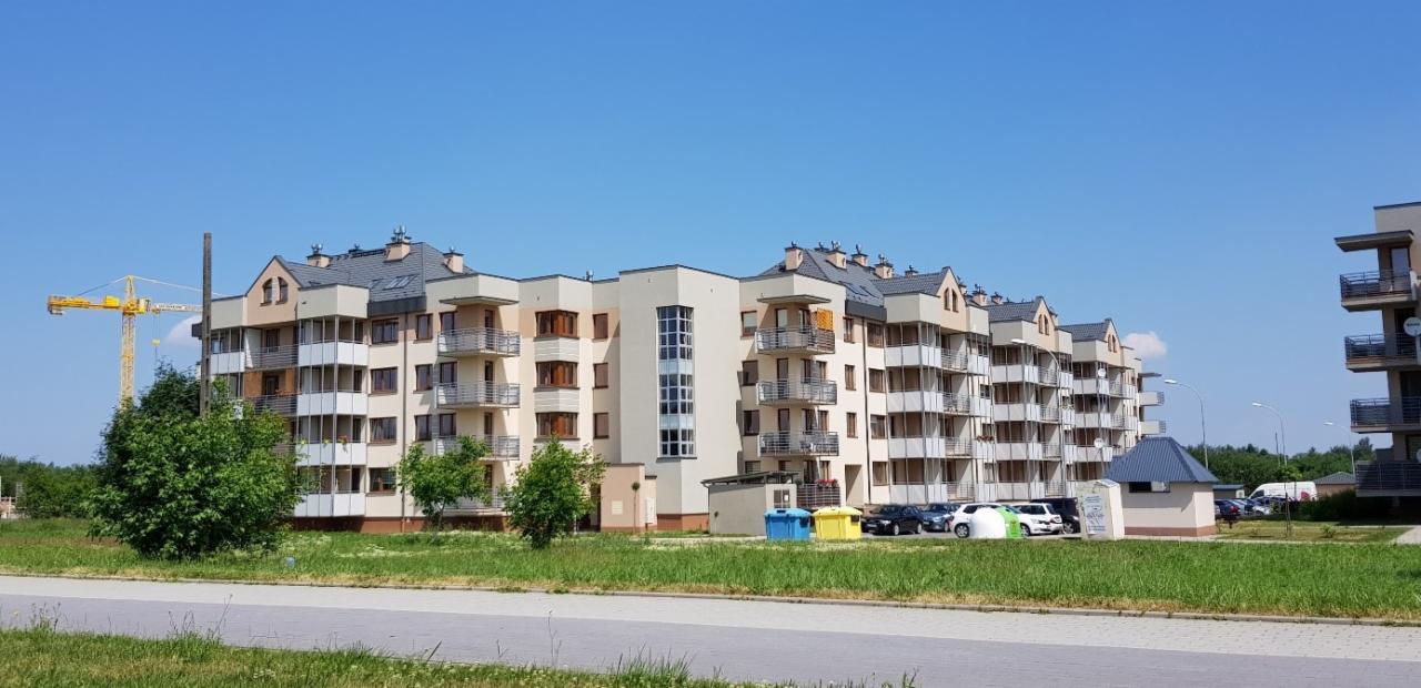 Nowoczesny, wielorodzinny budynek mieszkalny z balkonami, widoczny z poziomu ulicy, z żółtym żurawiem budowlanym w tle po lewej stronie.