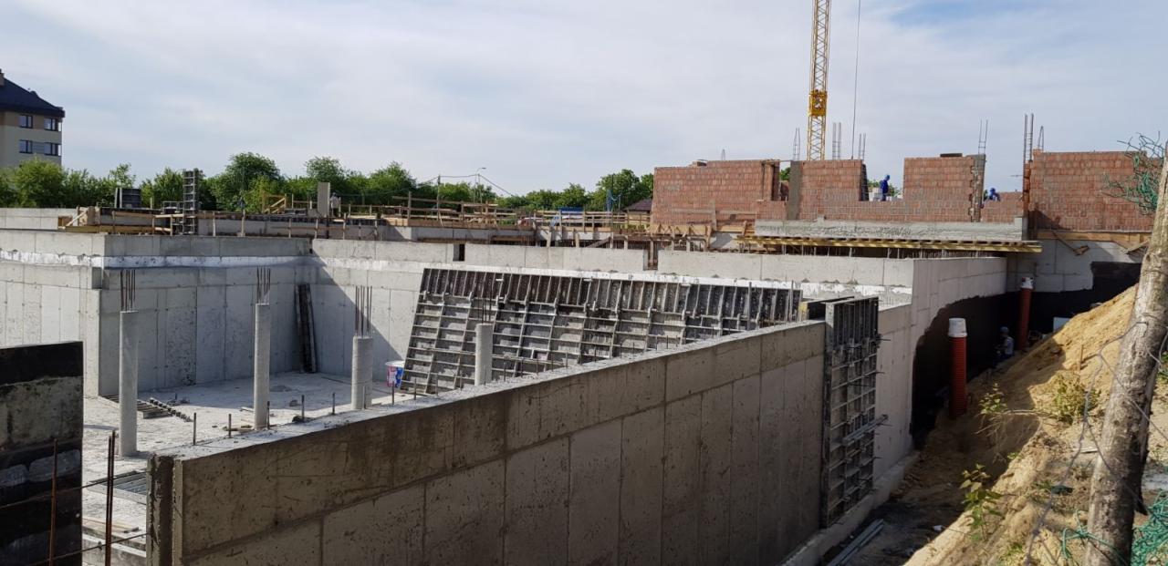 Fundamenty domu w trakcie budowy, widoczne betonowe ściany szalunkowe, zbrojenia, konstrukcja murowana w tle oraz żuraw budowlany.