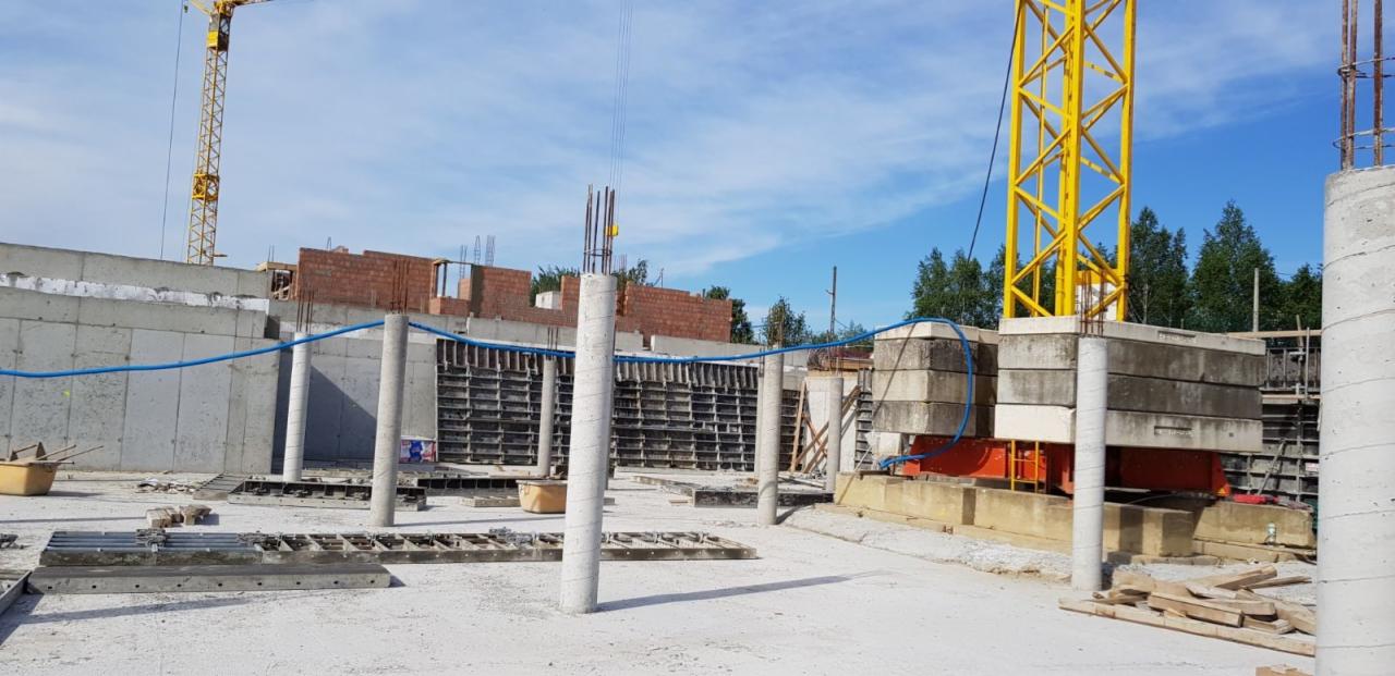 Fundamenty pod budynek w trakcie wylewania, widoczne szalunki, zbrojenia i betonowe kolumny, w tle fragment muru z cegły i żuraw budowlany.