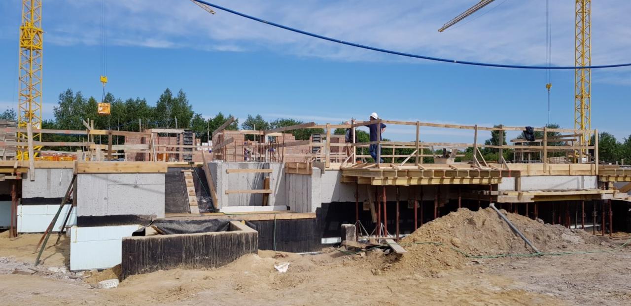 Budowa domu w trakcie realizacji: widoczne fundamenty z izolacją, szalunki, dźwigi w tle i pracownik w kasku.