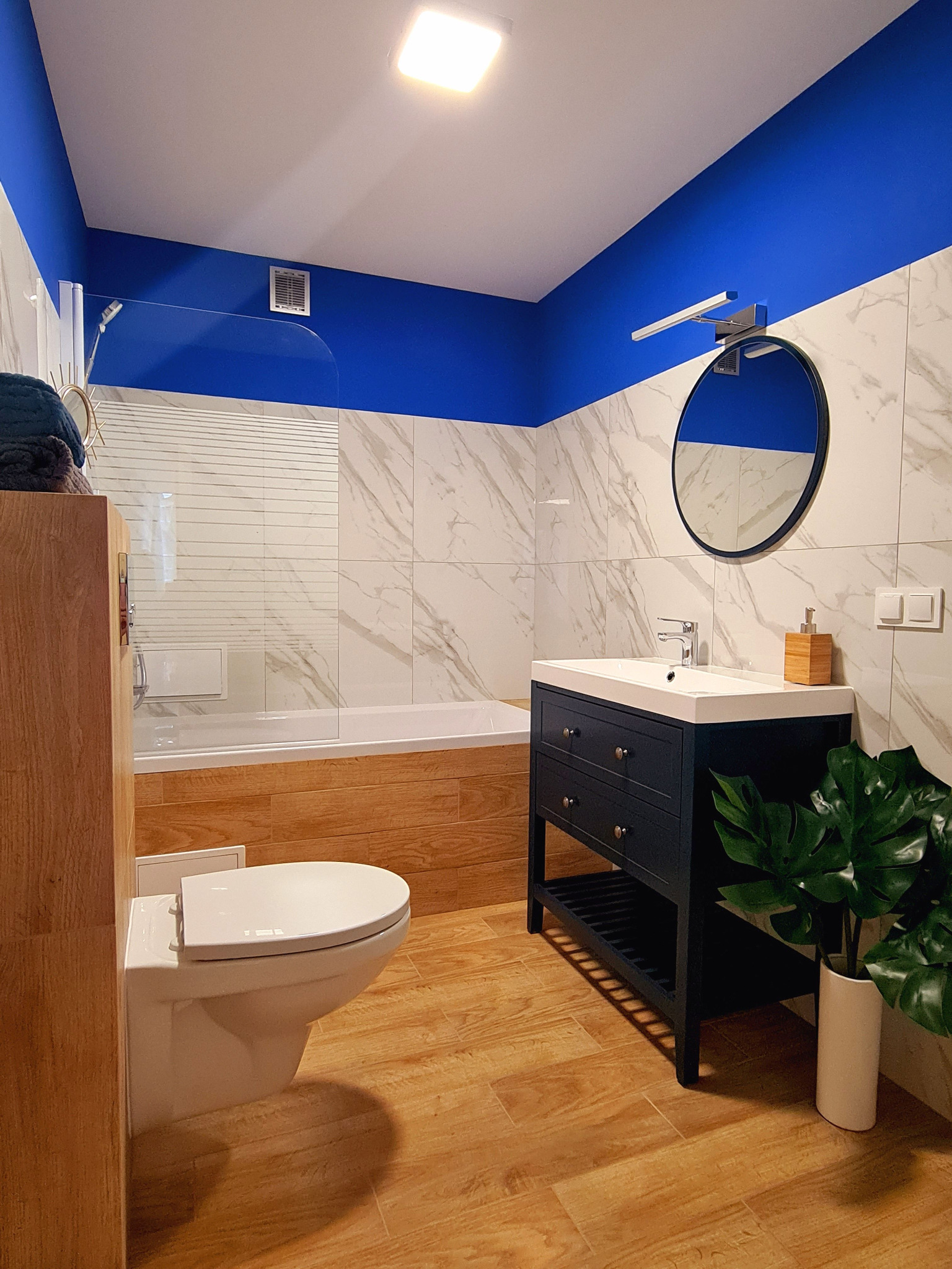 Nowoczesna łazienka z drewnopodobną podłogą i okładziną w strefie wanny, biało-marmurowymi płytkami ściennymi, umywalką z szafką w kolorze granatowym, okrągłym lustrem i akcentem w postaci...