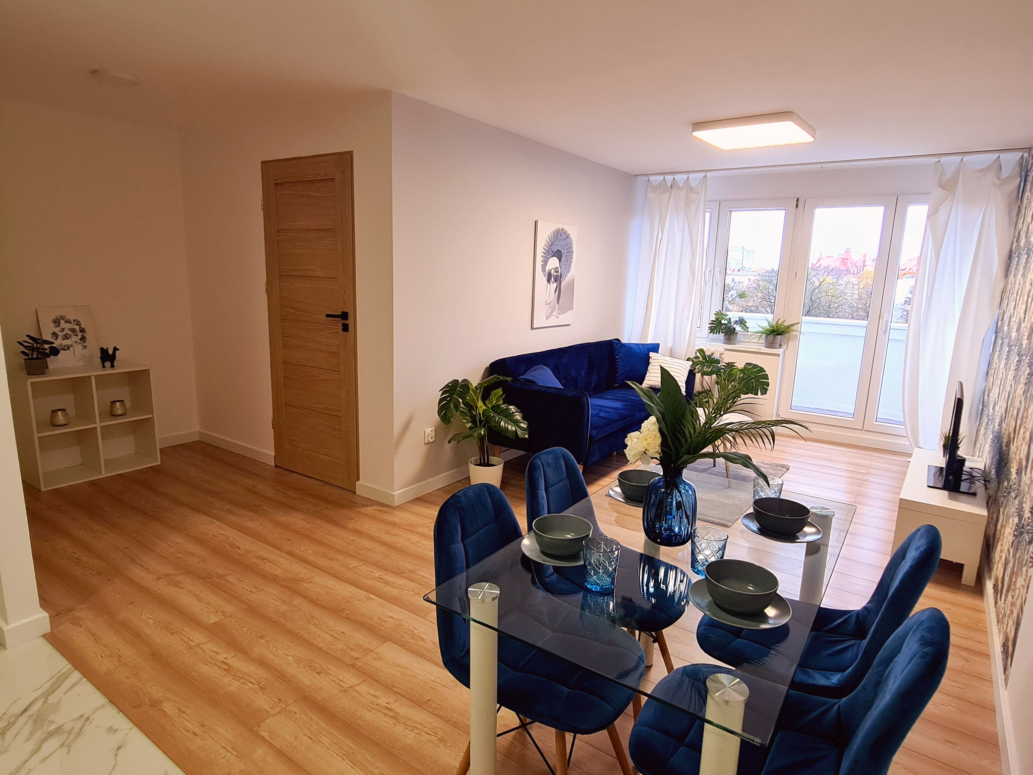 Jasne wnętrze mieszkania z drewnianą podłogą, szklanym stołem z aksamitnymi krzesłami w kolorze granatowym, granatową sofą i białymi zasłonami w oknach balkonowych.