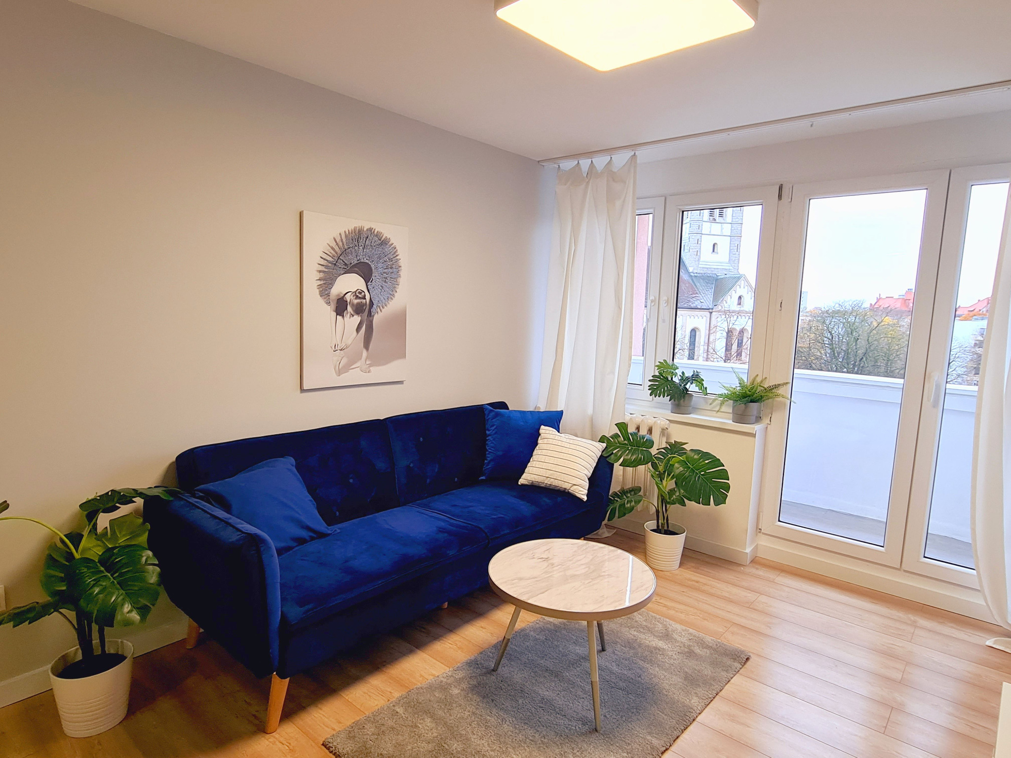 Salon z granatową sofą z weluru, okrągłym stolikiem kawowym z marmurowym blatem, dywanem i roślinami w doniczkach, widok na kościół za oknem.