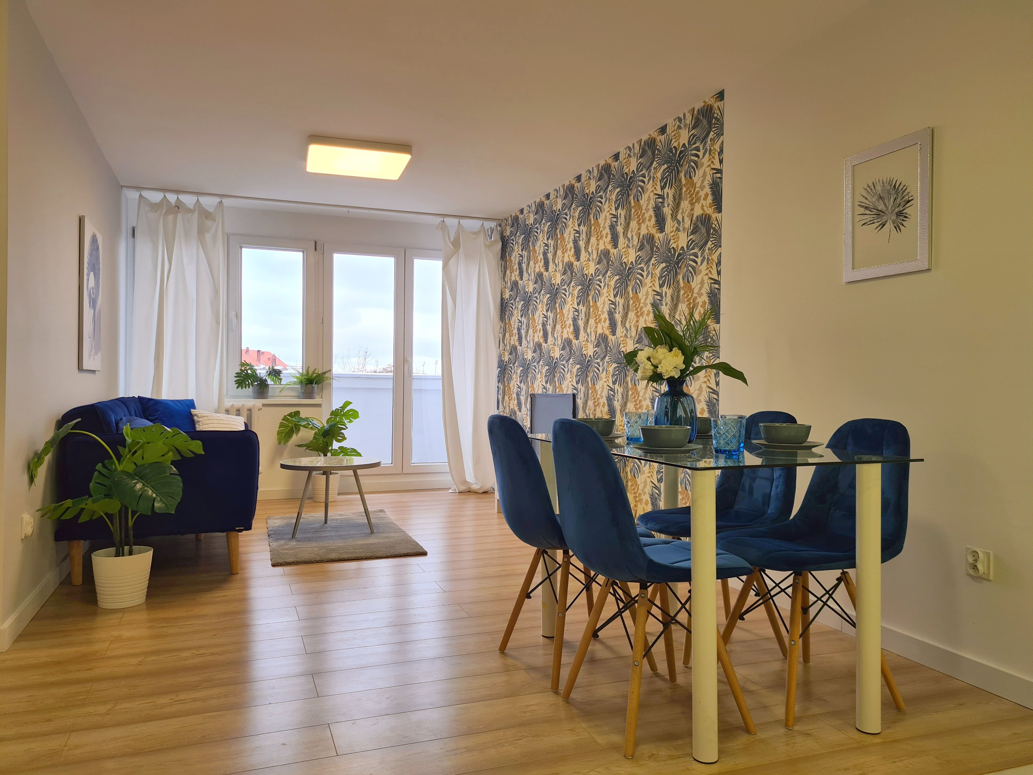 Jasne wnętrze mieszkania z niebieską sofą, szklanym stołem jadalnym z aksamitnymi krzesłami, tapetą w tropikalne liście i roślinami doniczkowymi, wykończone w nowoczesnym stylu.