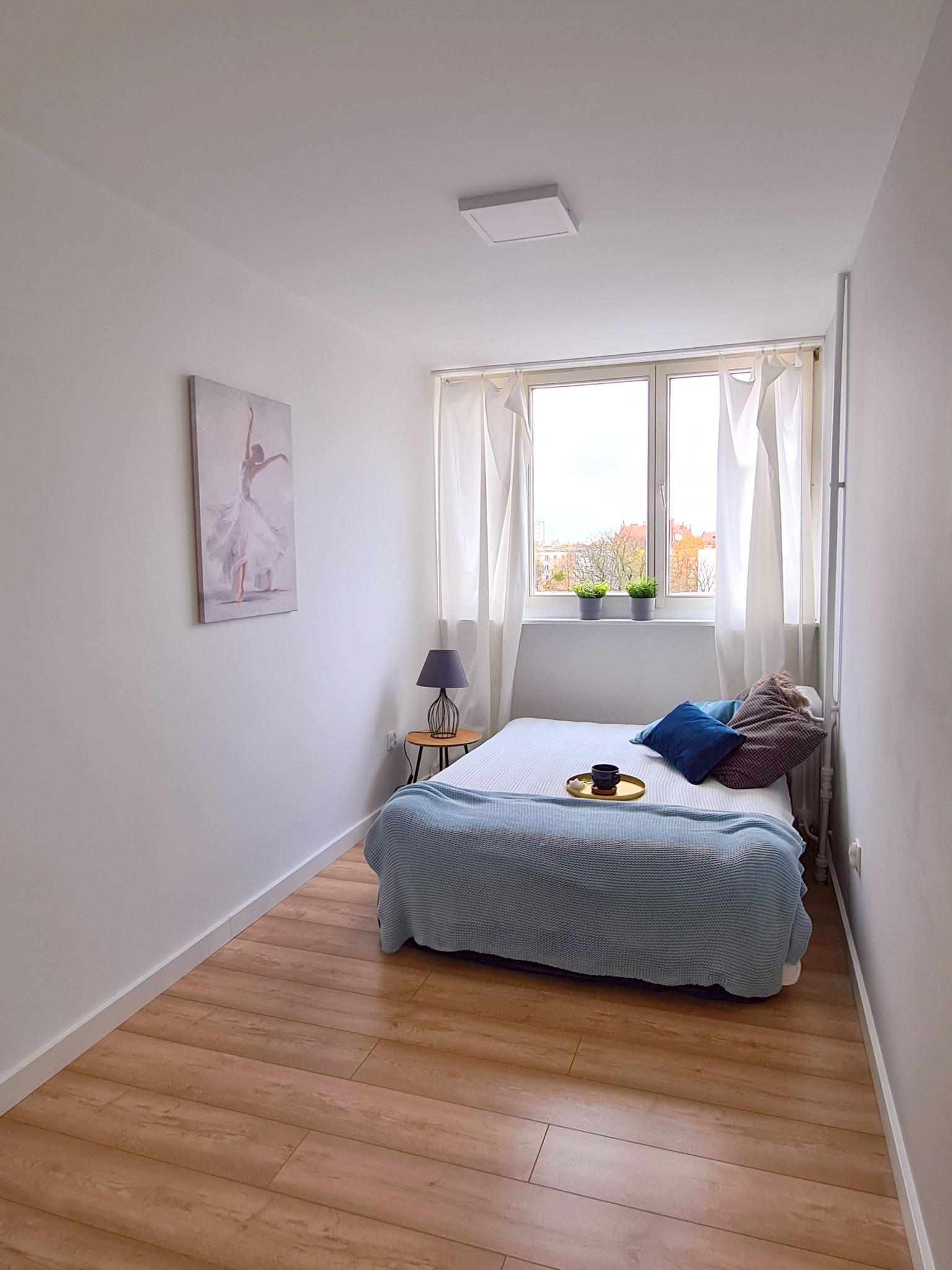 Mała sypialnia z łóżkiem, drewnianą podłogą i obrazem baletnicy na ścianie, w tle okno z roślinami.