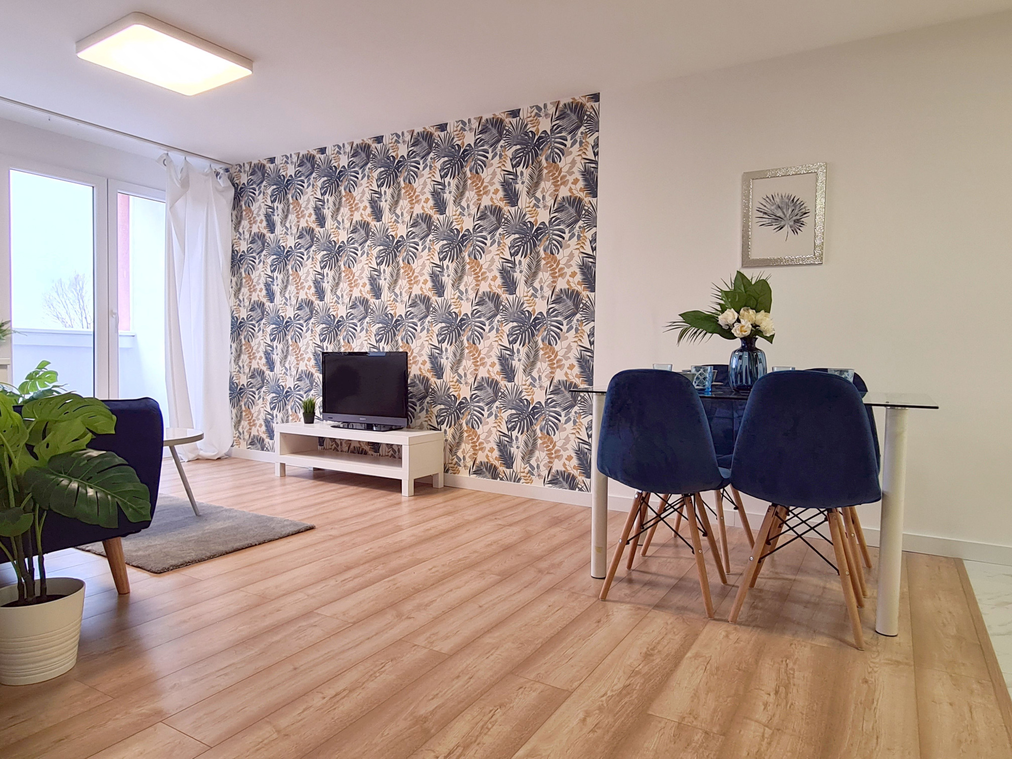 Jasne wnętrze salonu z drewnianą podłogą, ścianą z tapetą w motywy roślinne, telewizorem na białej szafce RTV, niebieskimi krzesłami welurowymi przy szklanym stole i rośliną doniczkową w białej...