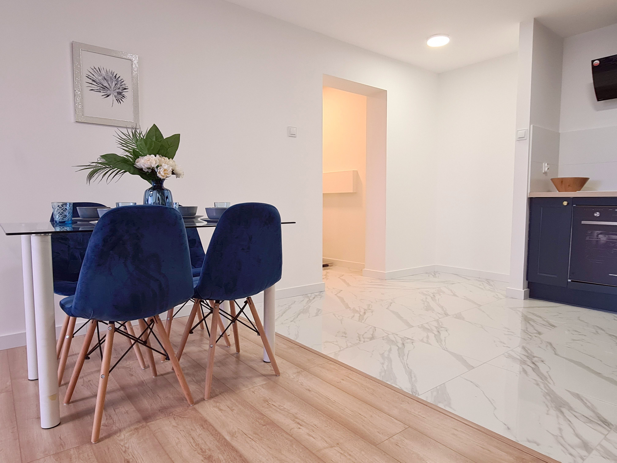Nowoczesna jadalnia z szklanym stołem, aksamitnymi krzesłami w kolorze granatowym, marmurową podłogą i drewnianą podłogą w strefie jadalnej, dekoracją kwiatową i minimalistycznym obrazem na ścianie.