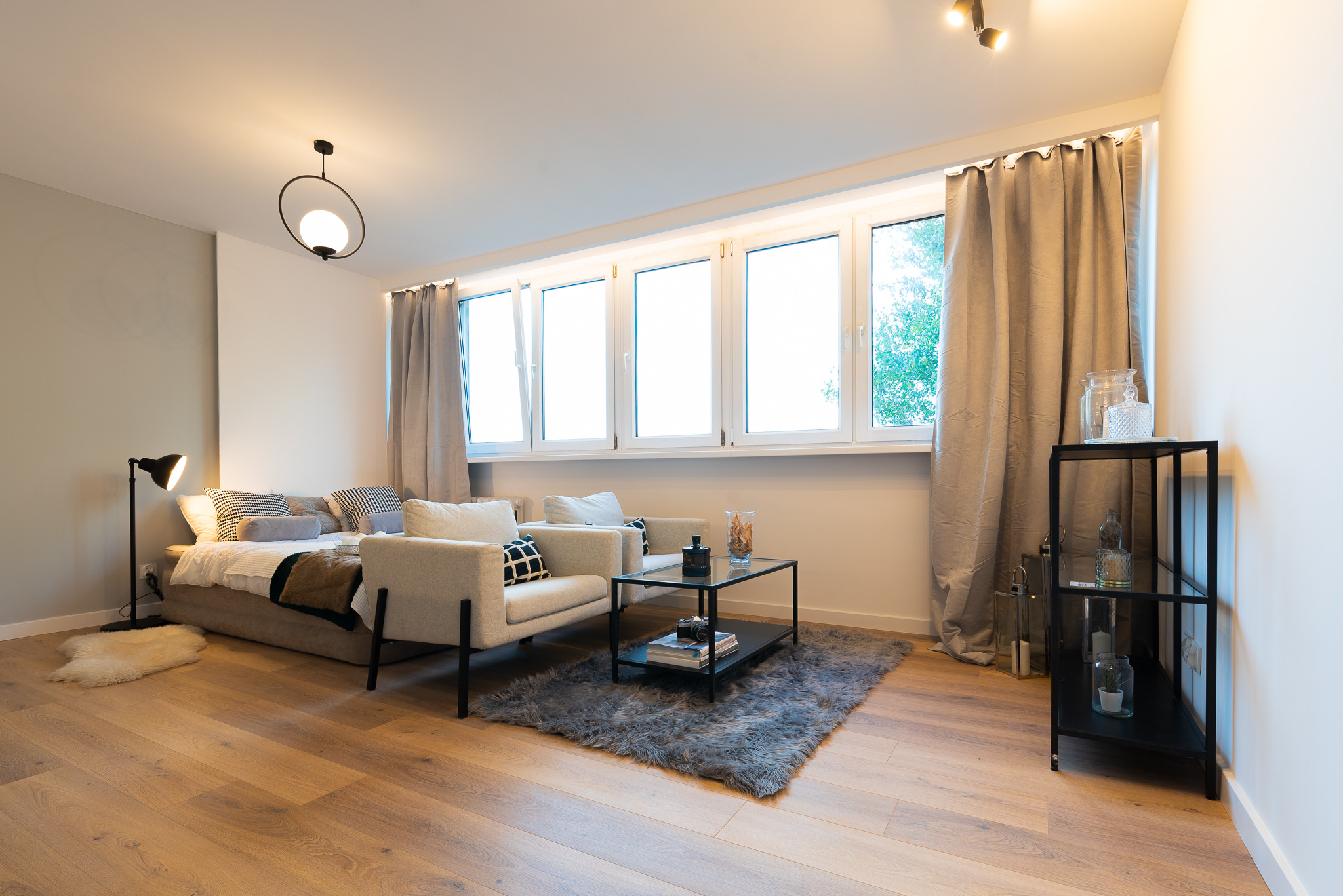 Jasne wnętrze pokoju dziennego z drewnianą podłogą, łóżkiem pełniącym funkcję sofy, dwoma fotelami, stolikiem kawowym na szarym dywanie i czarnym regałem pod oknem.