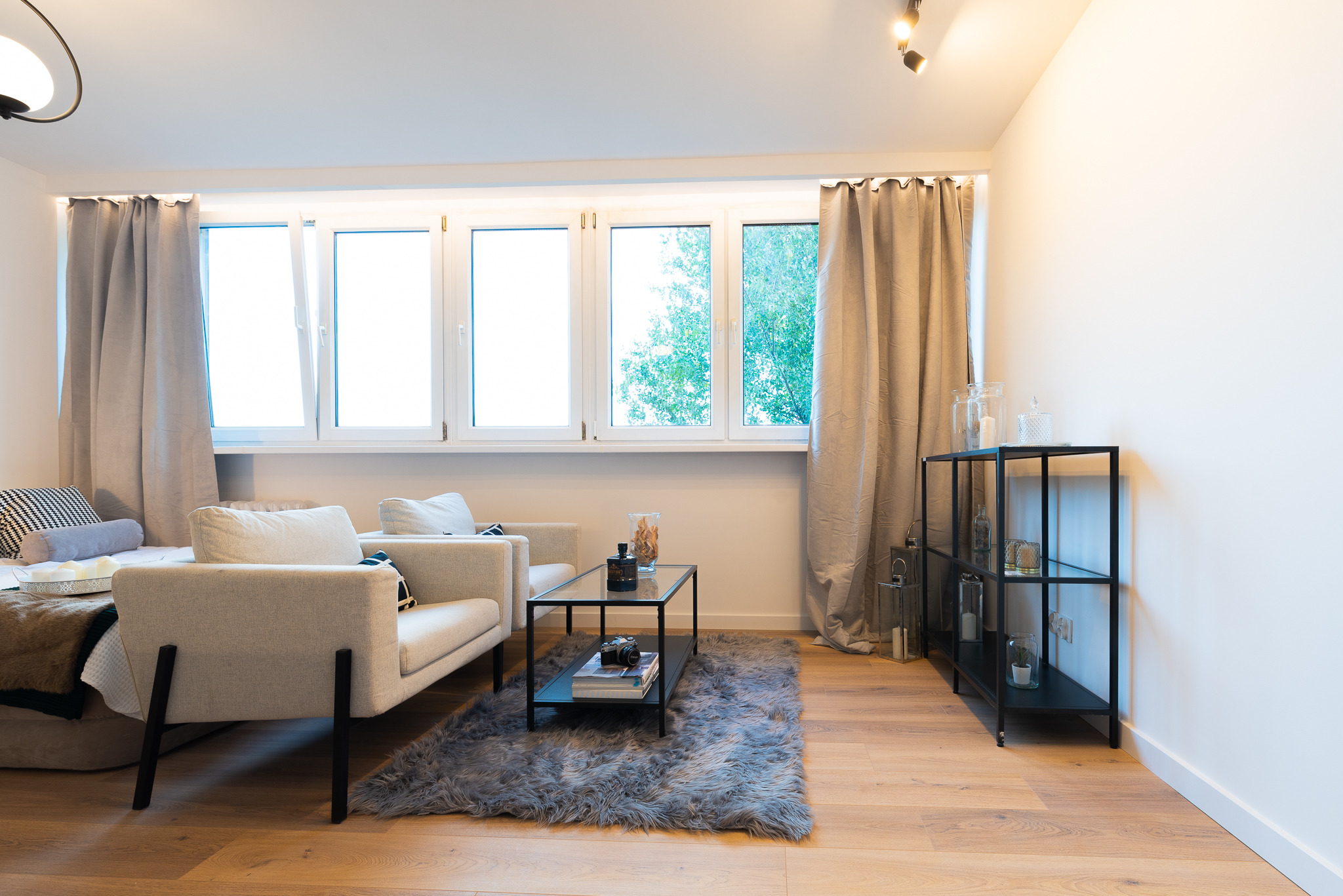 Jasne wnętrze salonu z drewnianą podłogą, szarym dywanem typu shaggy, dwoma beżowymi fotelami, czarnym stolikiem kawowym i regałem, okno z zasłonami w kolorze taupe.