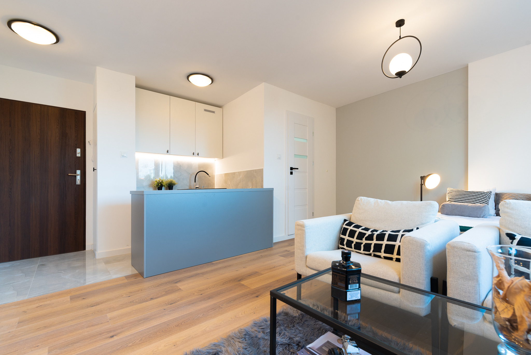 Nowoczesne wnętrze mieszkania we Wrocławiu: jasny salon z aneksem kuchennym, drewniana podłoga, szara wyspa kuchenna, białe szafki, minimalistyczne oświetlenie, biała sofa z geometrycznymi...