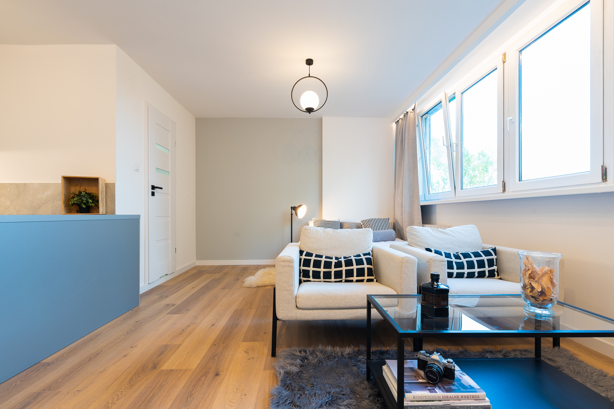 Jasne, nowocześnie urządzone wnętrze salonu z drewnianą podłogą, dwoma kremowymi fotelami z czarno-białymi poduszkami, stolikiem kawowym z czarną ramą i szklanym blatem, oraz dekoracjami: lampą...