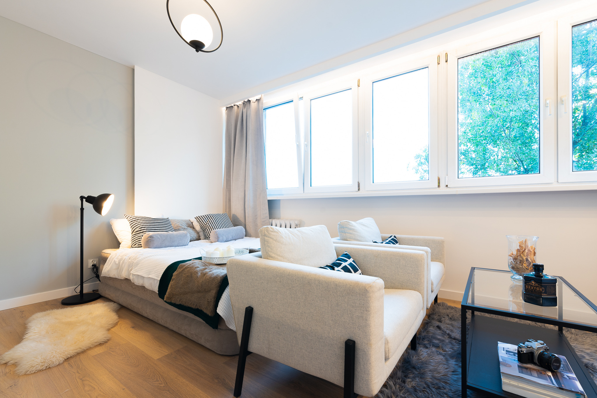 Nowoczesna aranżacja sypialni z łóżkiem na podeście, dwoma fotelami w jasnej tapicerce, stolikiem kawowym z dekoracjami i widokiem z okna na zieleń