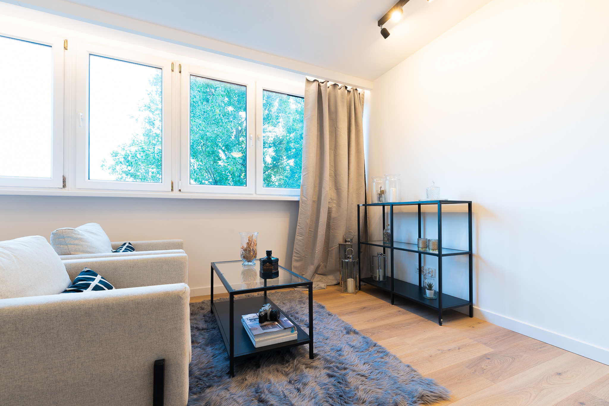 Jasny salon z drewnianą podłogą, dwoma fotelami, szarym dywanem imitującym futro i czarnymi meblami, oświetlony światłem dziennym wpadającym przez okno.