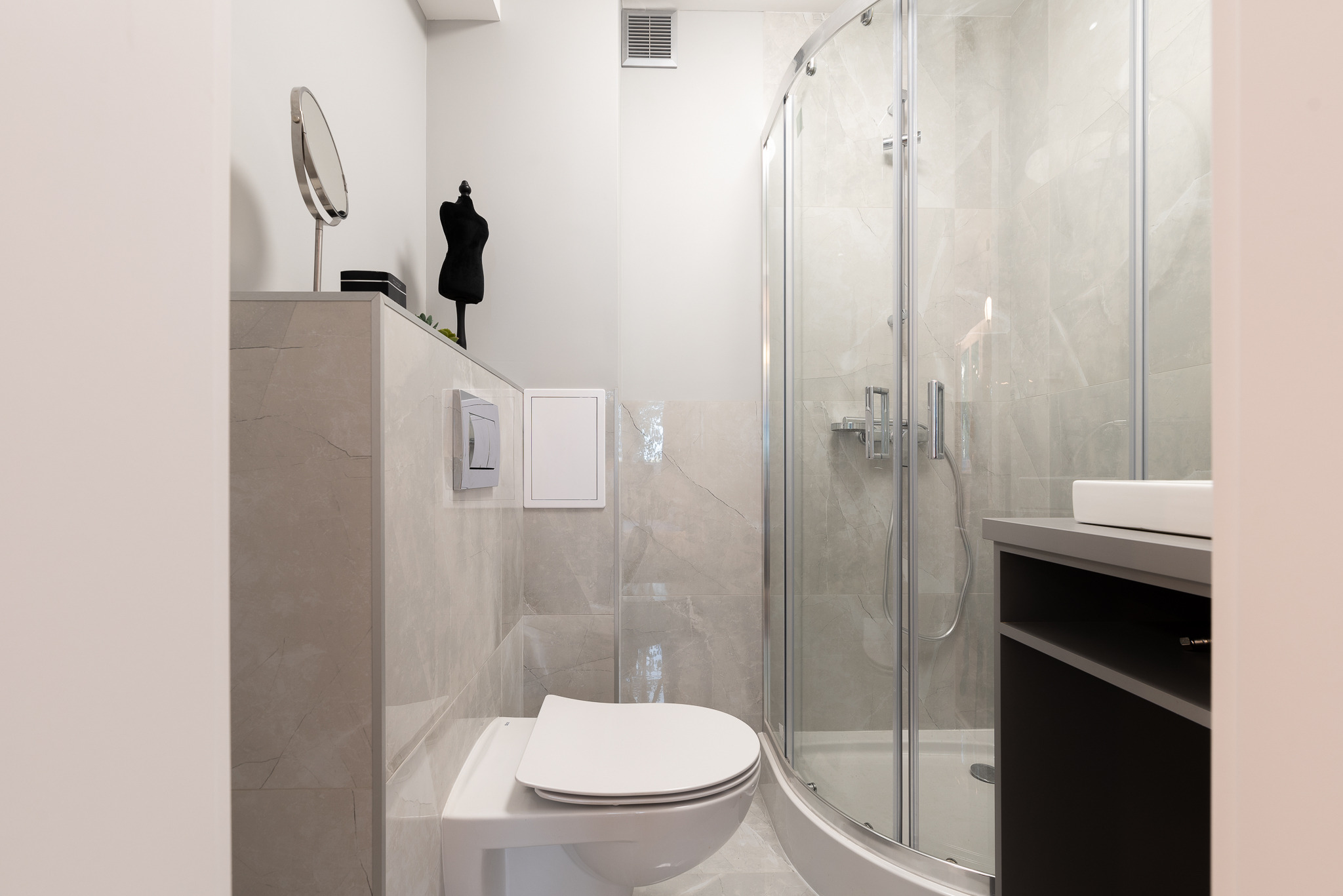 Nowoczesna łazienka z kabiną prysznicową, umywalką nablatową na szafce, toaletą wiszącą i dekoracyjnymi elementami: lustro na postumencie i czarna figurka na półce, wykończona płytkami imitującymi...