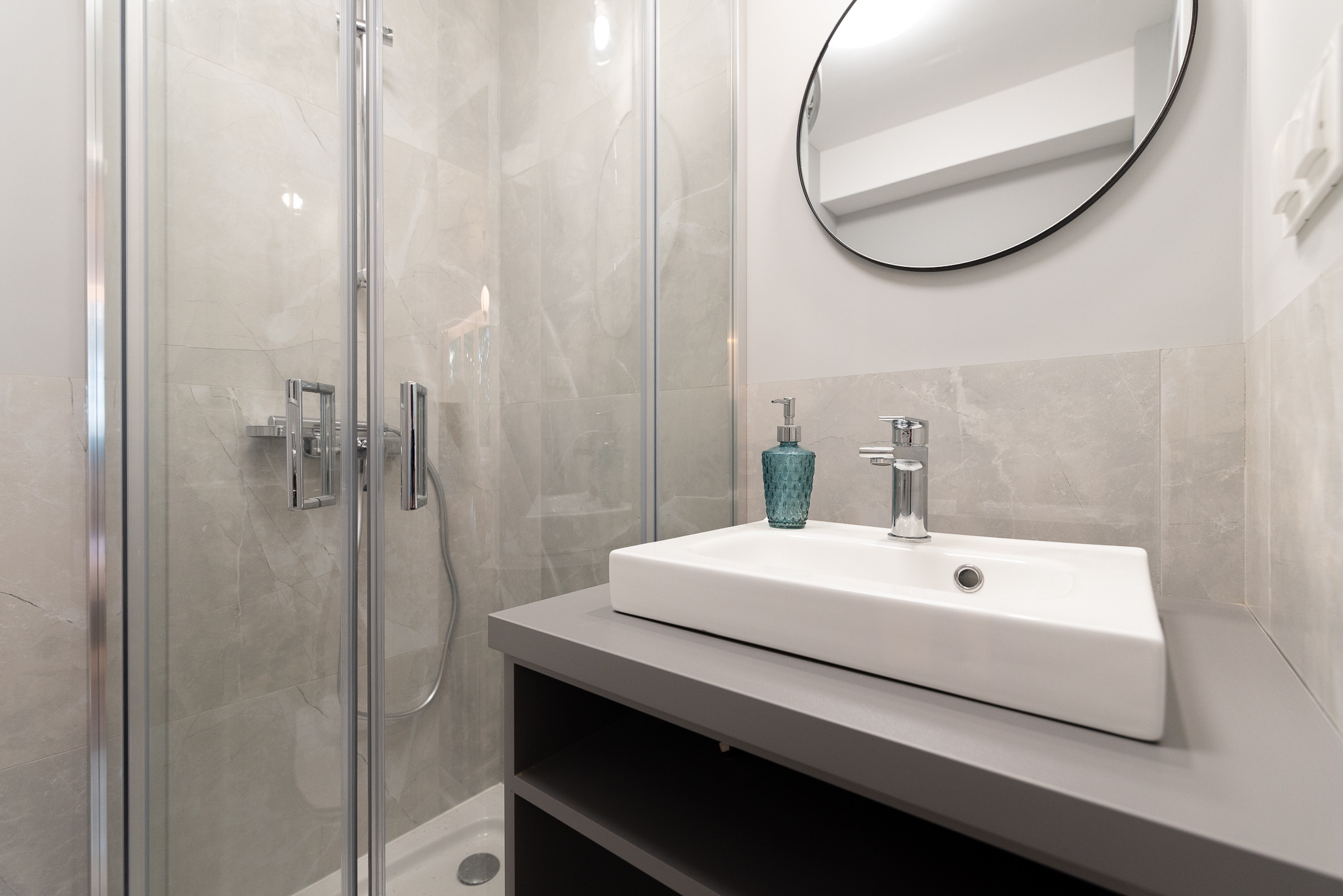 Nowoczesna łazienka z kabiną prysznicową, umywalką nablatową na szarej szafce i okrągłym lustrem, wykończona płytkami imitującymi marmur