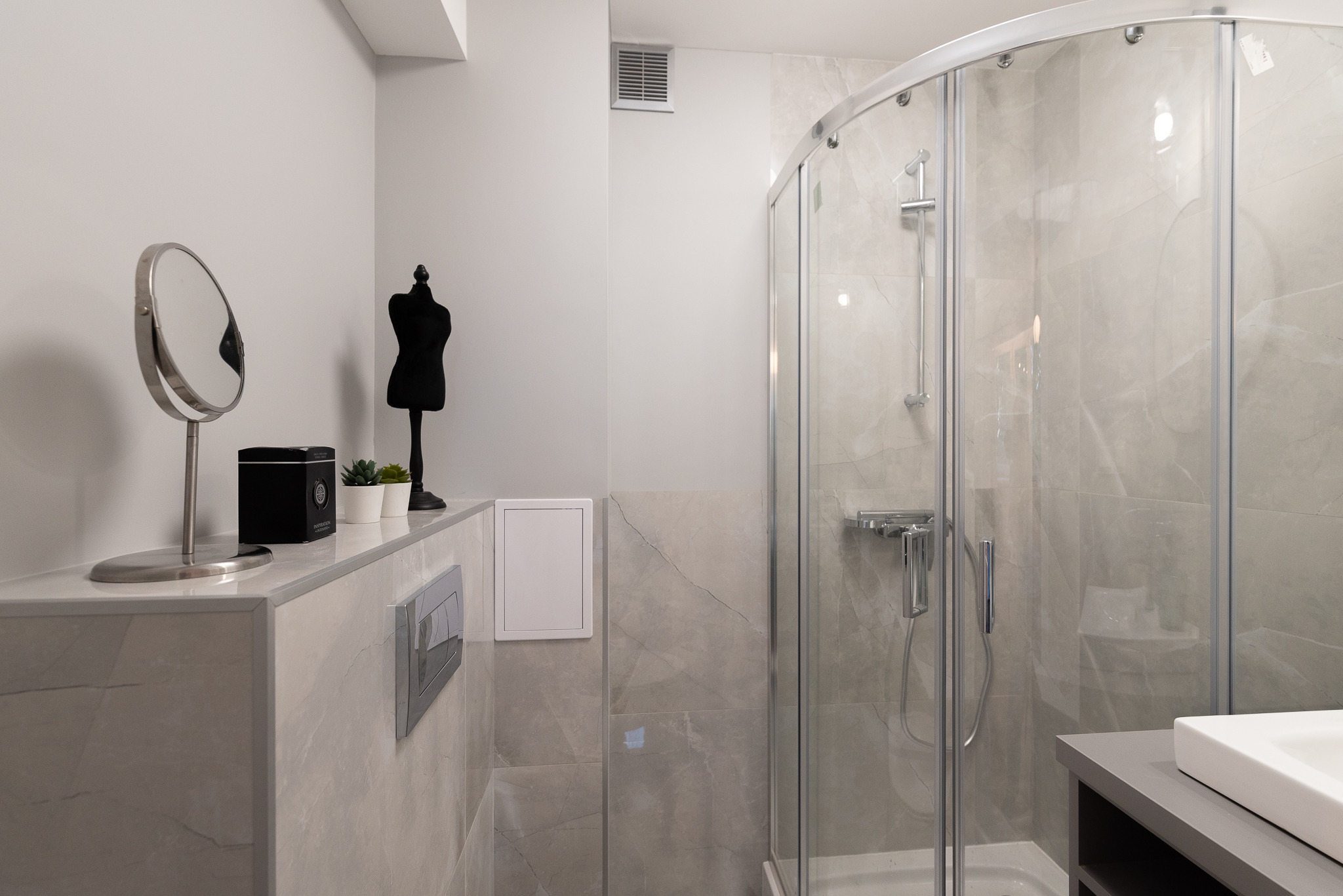 Nowoczesna łazienka w odcieniach szarości z kabiną prysznicową, lustrem na metalowej podstawie, czarną figurką manekina, małymi roślinami w białych doniczkach i ukrytą spłuczką WC.