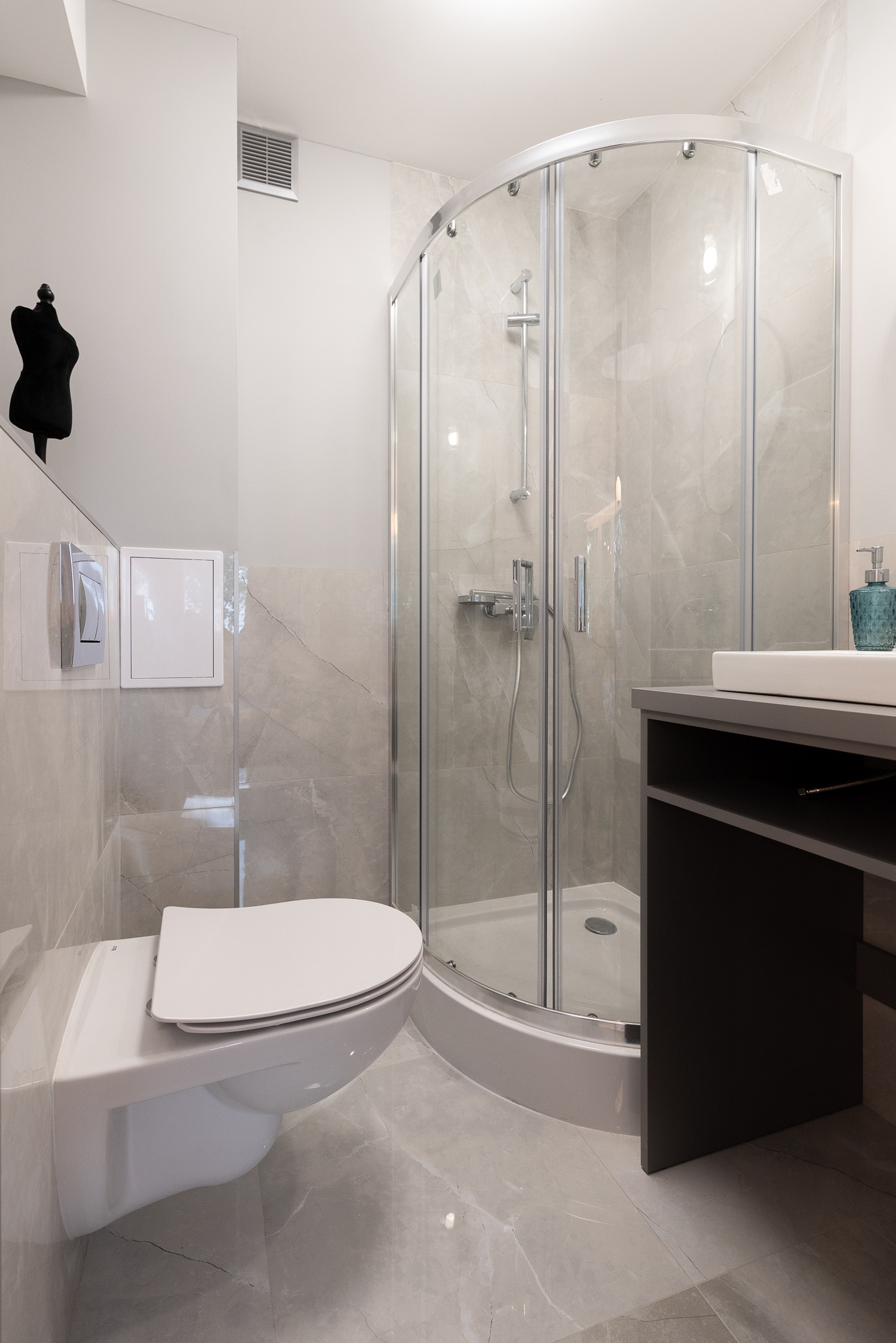 Nowoczesna łazienka z kabiną prysznicową, podwieszaną toaletą i umywalką nablatową na ciemnoszarym blacie. Szare płytki imitujące marmur na podłodze i ścianach. W tle, na ścianie, dekoracyjna...