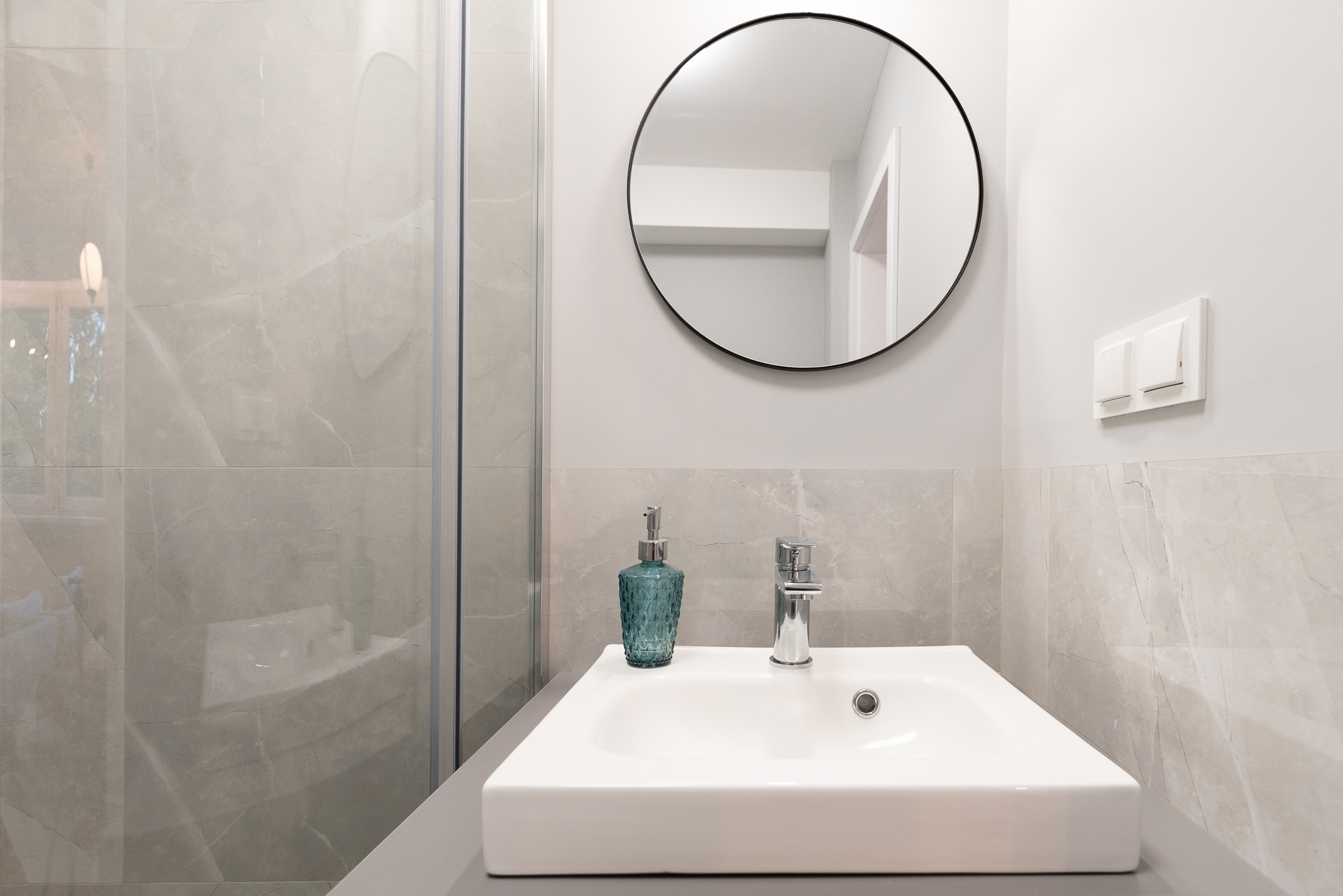 Nowoczesna łazienka z umywalką nablatową, chromowaną baterią, dekoracyjnym dozownikiem mydła w odcieniach turkusu, okrągłym lustrem w czarnej ramie i kabiną prysznicową wyłożoną płytkami...