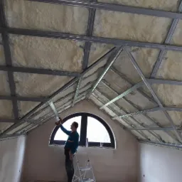 Pracownik w trakcie montażu metalowej konstrukcji sufitu podwieszanego z ociepleniem wełną mineralną na poddaszu, widoczna drabina i okno łukowe.