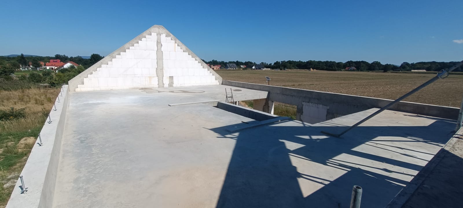 Surowy stan dachu wznoszonego domu jednorodzinnego z białych bloczków, widok z góry na rozległe pola w tle, na dachu drabina i wystające pręty zbrojeniowe.