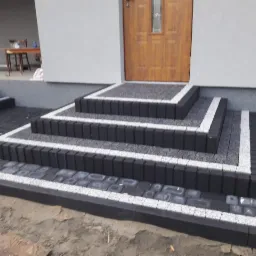 Schody wejściowe do domu wyłożone kostką brukową w odcieniach szarości i bieli, z obrzeżami z ciemnych kostek, prowadzące do drewnianych drzwi wejściowych.