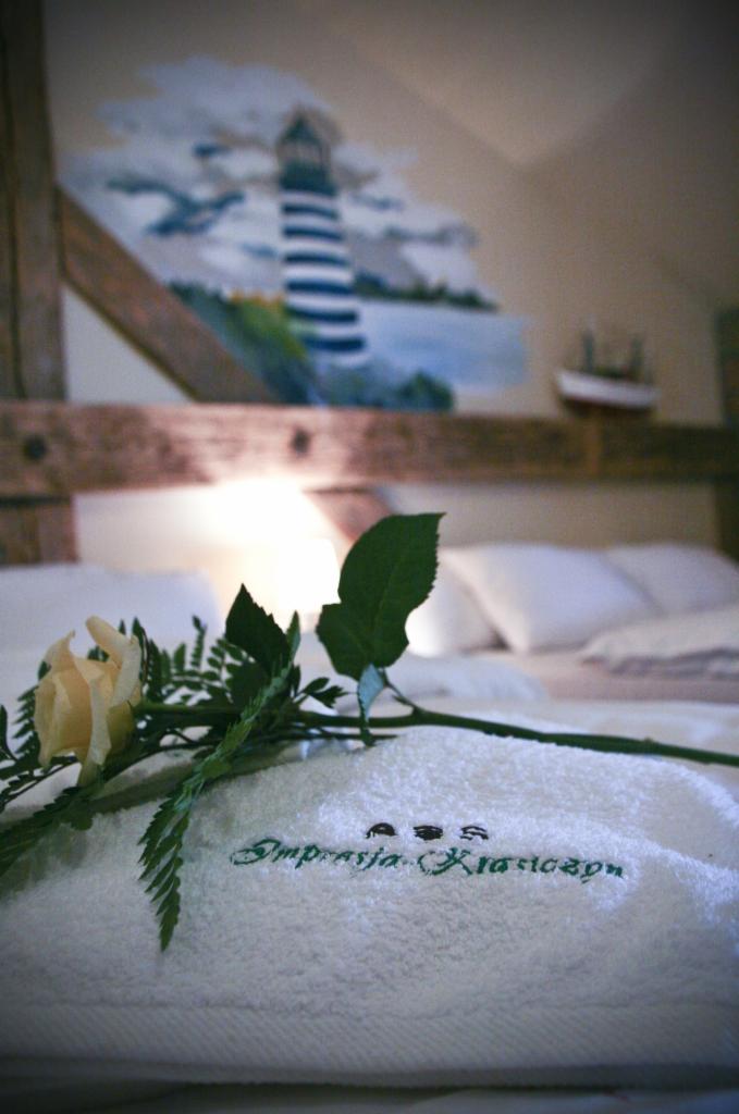 Udekorowany pokój hotelowy z ręcznikiem z haftem 'Impresja Klasyczna', różą i malowidłem latarni morskiej nad łóżkiem.