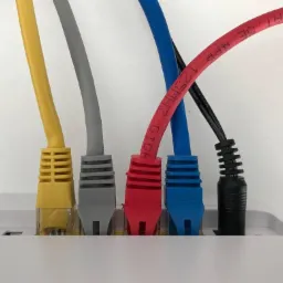 Pięć kolorowych kabli sieciowych (żółty, szary, czerwony, niebieski, czarny) podłączonych do białego routera. Widoczne oznaczenia na czerwonym kablu: 'TP CAT 5E 4PR 1.25MMHZ'.