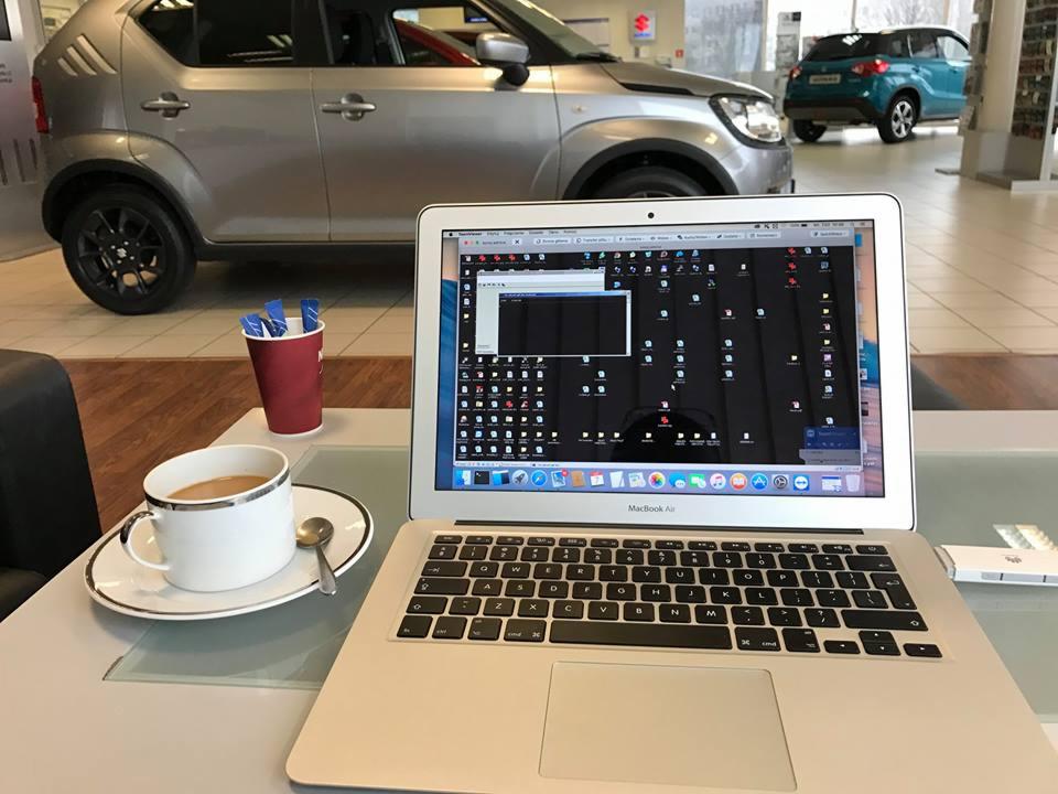 Laptop MacBook Air z otwartym ekranem, na którym widoczny jest pulpit z licznymi ikonami, stoi na jasnym biurku w salonie samochodowym Suzuki, obok filiżanka kawy z mlekiem i kubek z patyczkami...