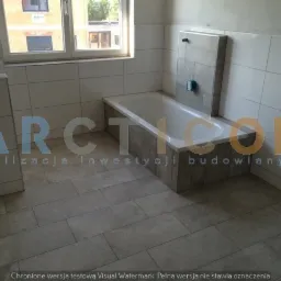 Wykończona łazienka w jasnych płytkach z białą wanną obudowaną płytkami imitującymi kamień, widoczne okno i fragment budynku za nim, nałożony znak wodny.