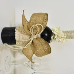 Udekorowana butelka wina, owinięta jutowym sznurkiem i koronką, z kokardą z juty i kremowymi kwiatami z perłami, leżąca obok kieliszków do wina.