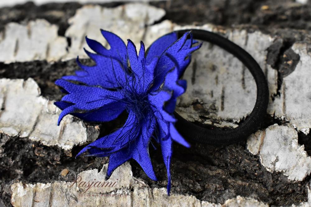 Niebieska gumka do włosów z materiałowym kwiatem na tle kory brzozy, z widocznym logo producenta w lewym dolnym rogu.