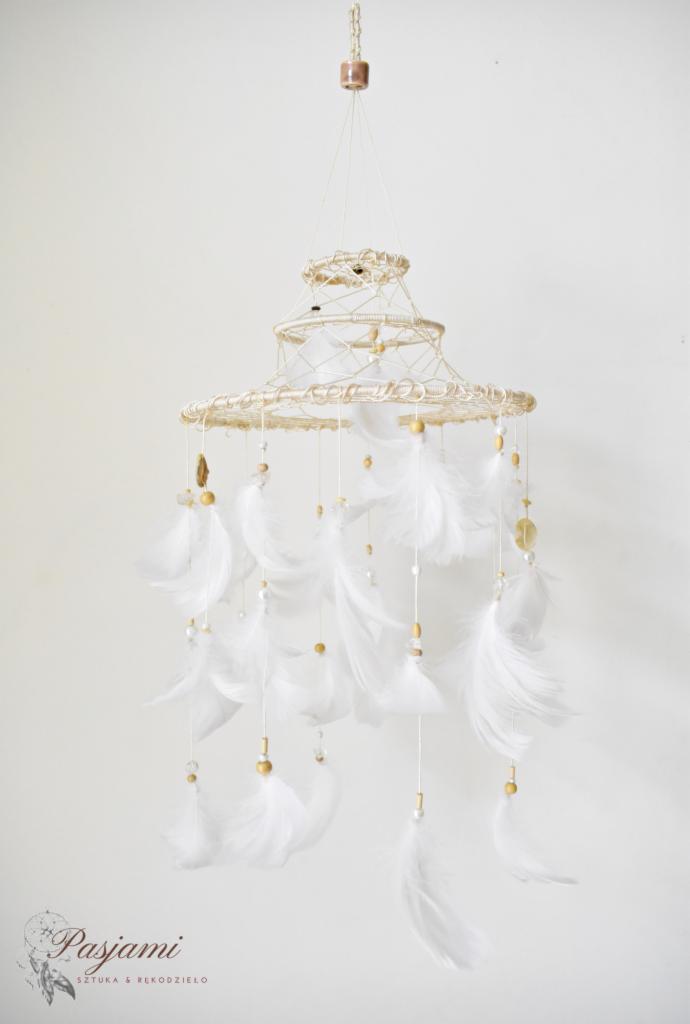 Ręcznie robiony, trójpoziomowy łapacz snów w odcieniach bieli i beżu, z piórami, drewnianymi koralikami i kryształkami, zawieszony na tle jasnej ściany.