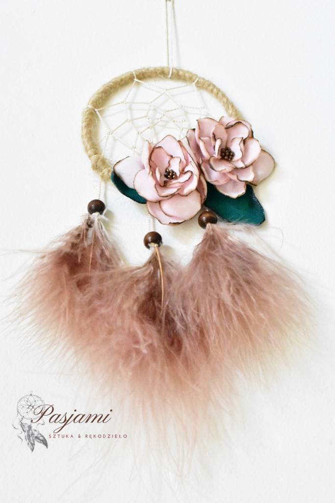 Ręcznie robiony łapacz snów z beżowym okręgiem, białymi nićmi, różowymi kwiatami z zielonymi liśćmi i zwisającymi różowymi piórami, zawieszony na białym tle; w lewym dolnym rogu logo firmy.