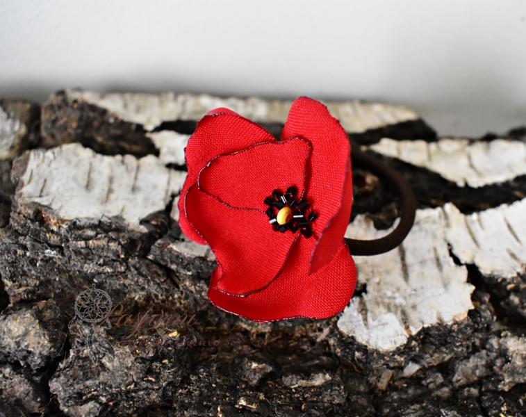 Czerwona gumka do włosów w kształcie maku z czarnymi koralikami, umieszczona na korze drzewa.