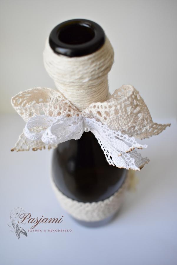 Dekoracyjna, ciemna butelka owinięta jutowym sznurkiem i ozdobiona koronkową kokardą z koralikiem, na białym tle.