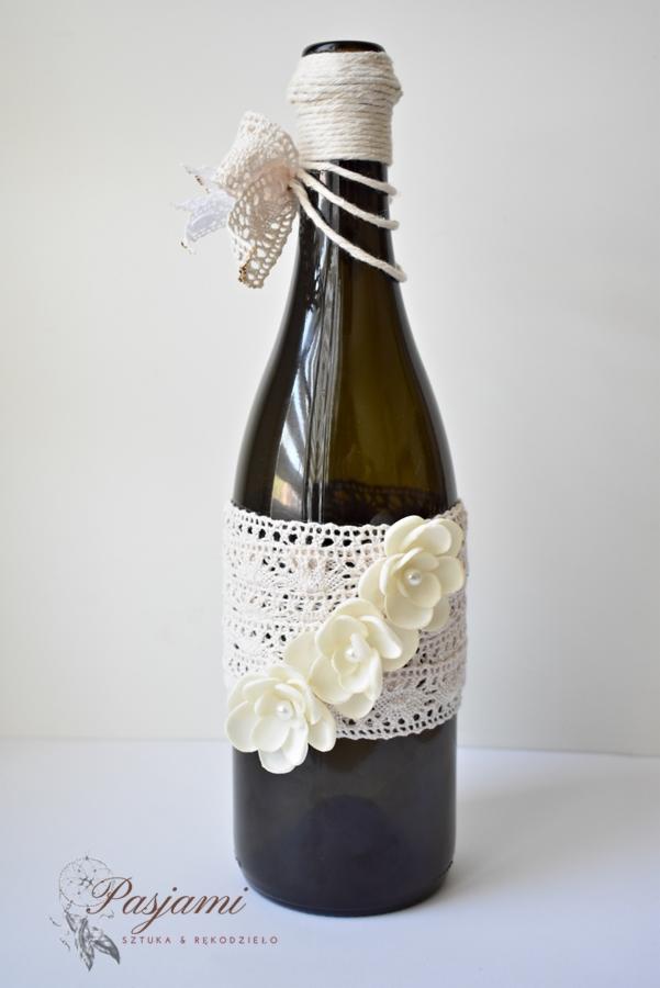 Dekoracyjna butelka z ciemnego szkła, owinięta koronkową wstążką z trzema białymi, perłowymi kwiatami, szyjka ozdobiona sznurkiem i koronkową kokardką.