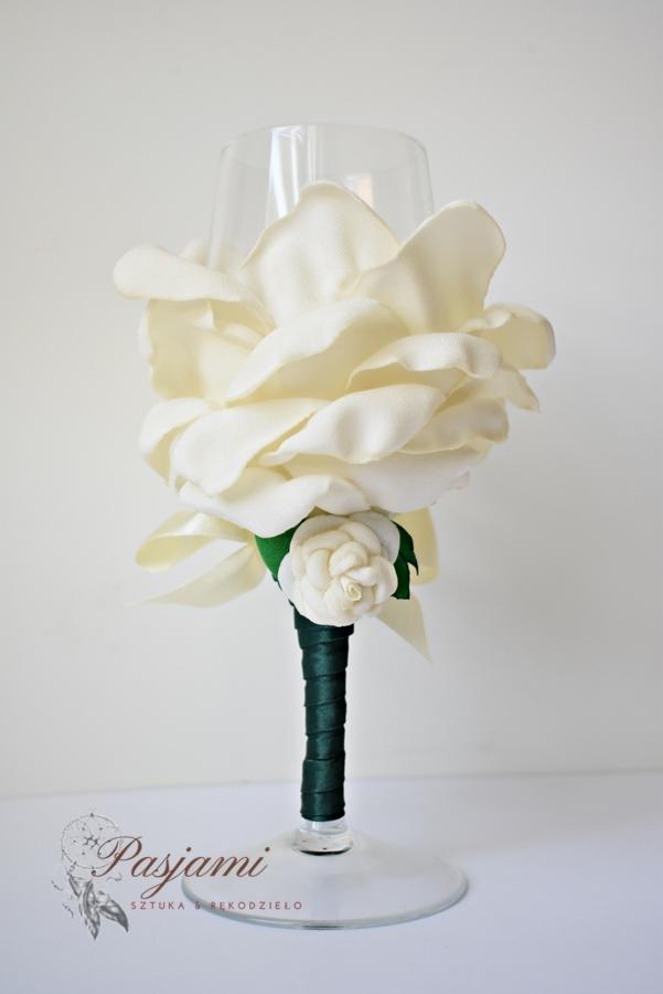 Udekorowany kieliszek do wina z dużą kremową różą z materiału i mniejszą kremową różą na zielonej łodydze z wstążki, na białym tle, z widocznym logo firmy Pasjami.